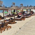 Un camarero, con visera blanca, sirve a unos turistas tumbados en una hamaca.