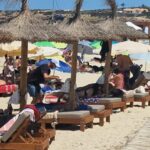 Un camarero, con visera blanca, sirve a unos turistas tumbados en una hamaca.