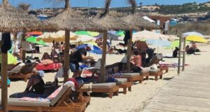 Un camarero, con visera blanca, sirve a unos turistas tumbados en una hamaca.