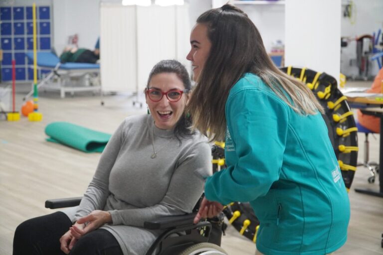Mujer en silla de ruedas sonríe mientras habla con una profesional en el gimnasio.