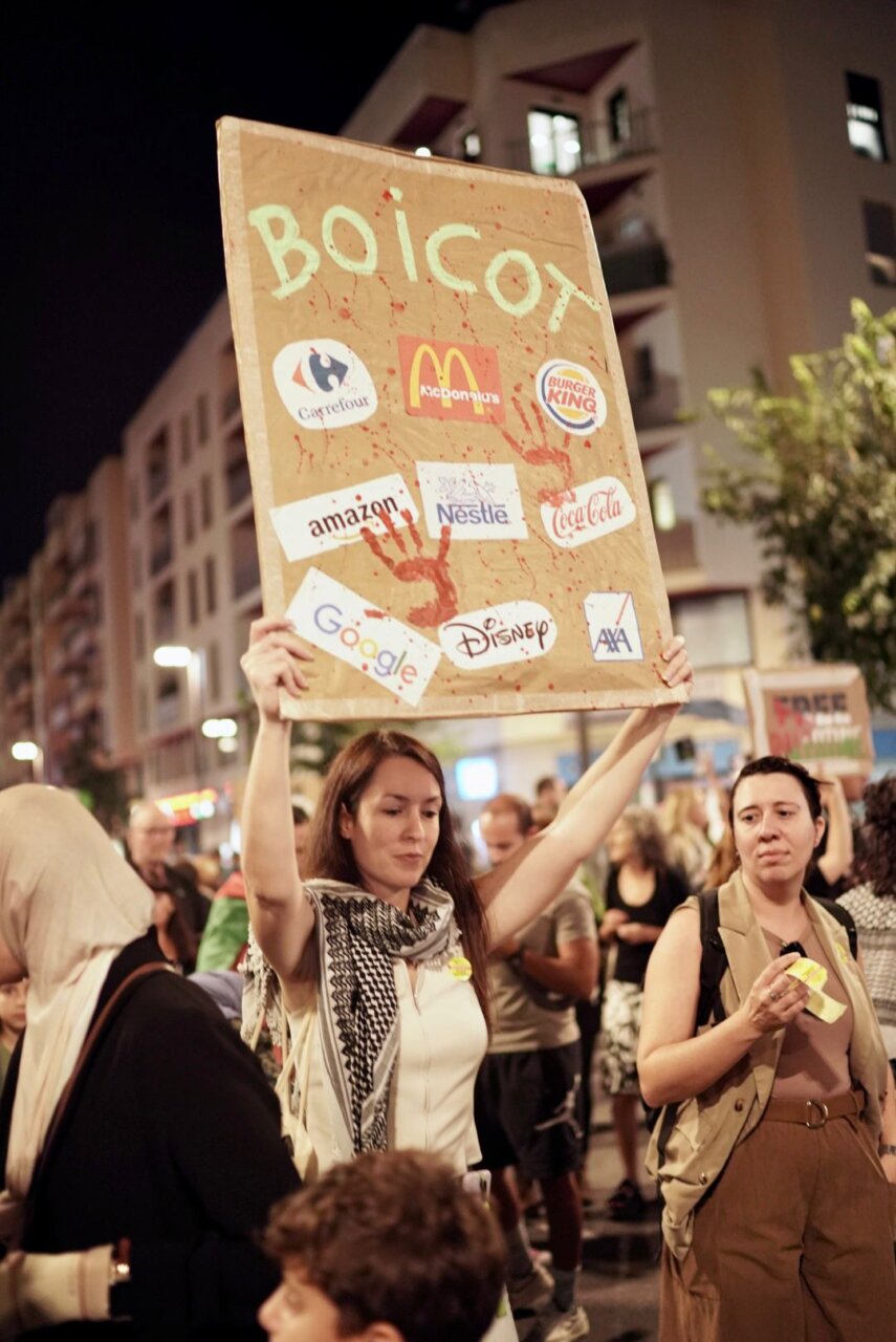 Persona sosteniendo un cartel de boicot en una manifestación nocturna.