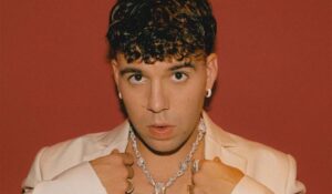 El artista Quevedo posando con una cadena de joyas en un fondo rojo.