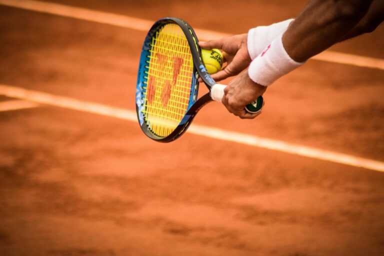 Mano sosteniendo una raqueta de tenis y una pelota en una cancha de tierra batida